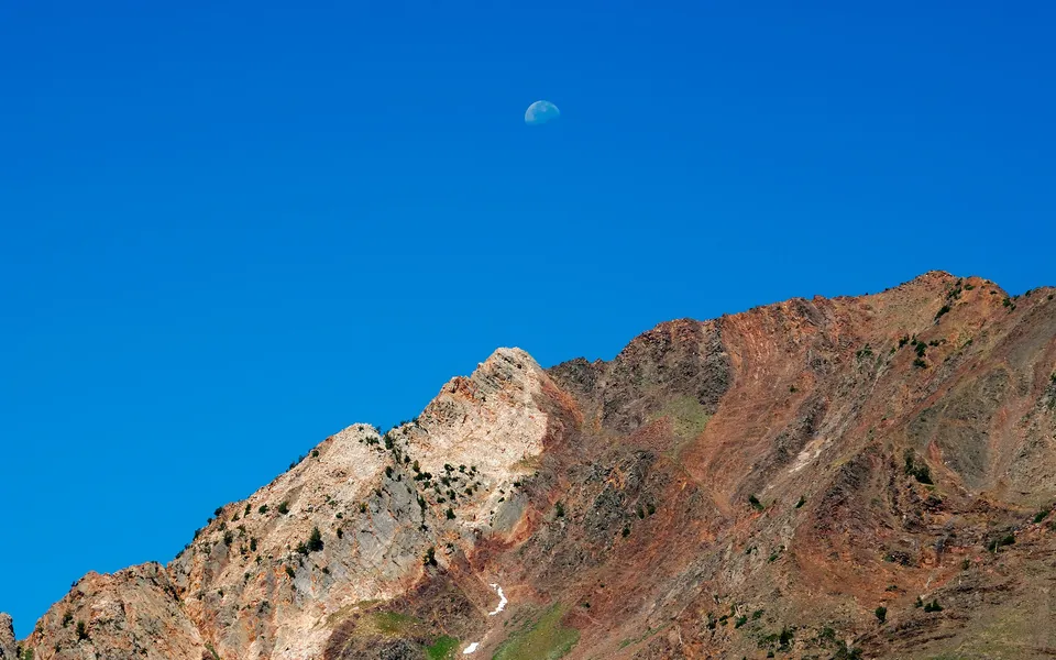 A rugged mountain range with layered rock formations under a clear blue sky with the moon visible.