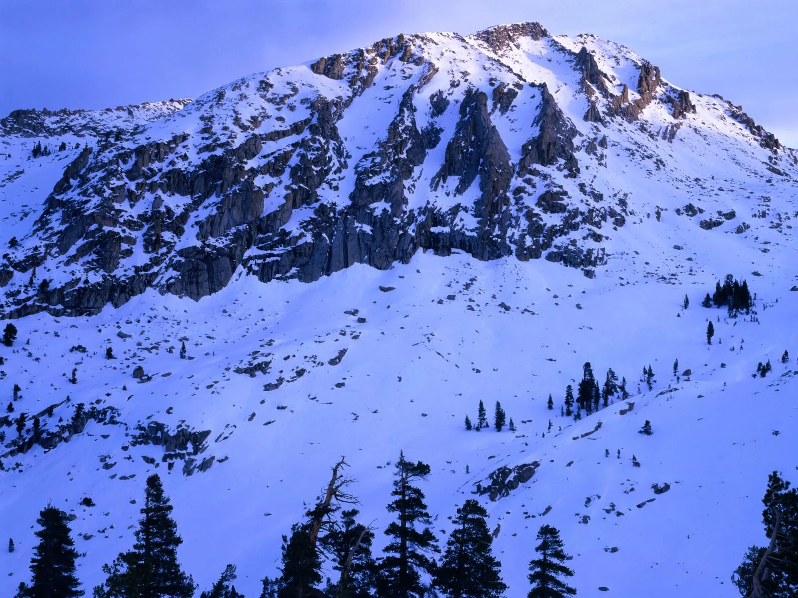 A rugged, snow-covered mountain slope is illuminated by soft, cool blue light, revealing rocky formations.