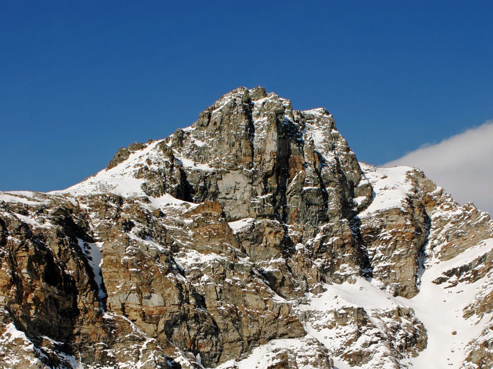 A rugged mountain summit with rocky faces partially covered in snow under a bright blue sky.