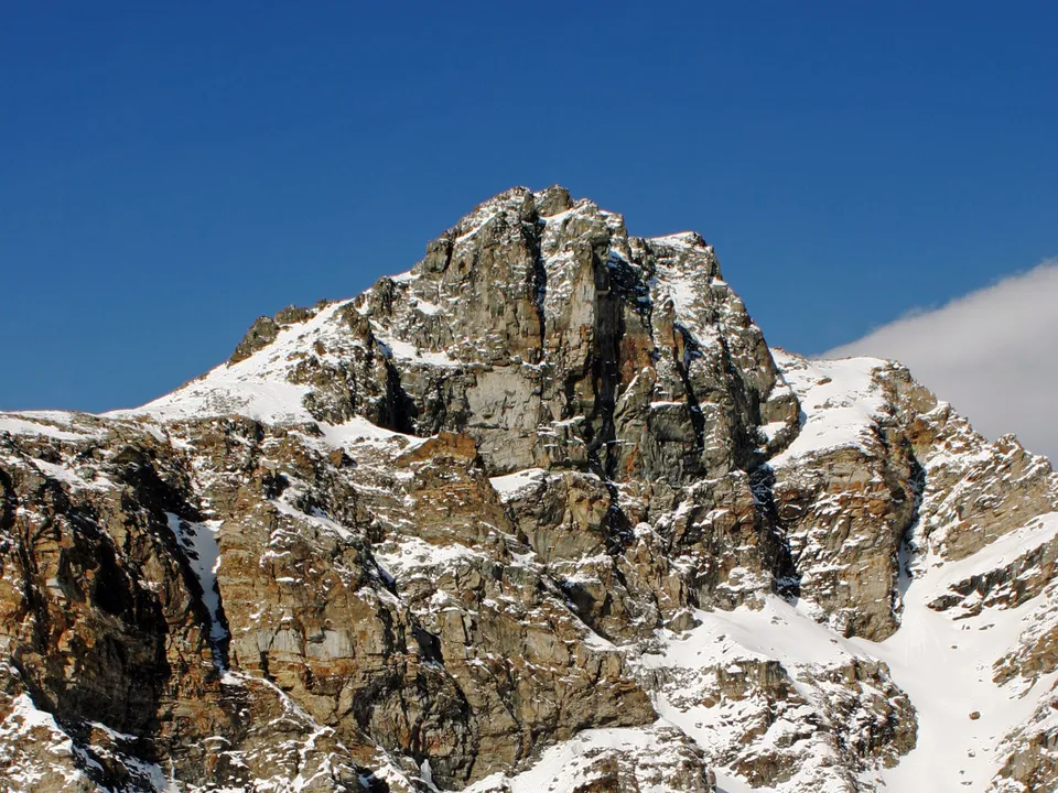 A rugged mountain summit with rocky faces partially covered in snow under a bright blue sky.
