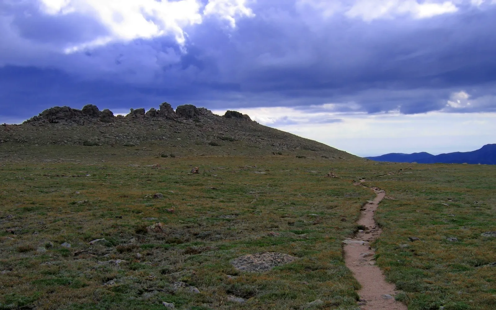 A dirt path winds through a grassy meadow towards a rocky mountain summit under a dramatic cloudy sky.