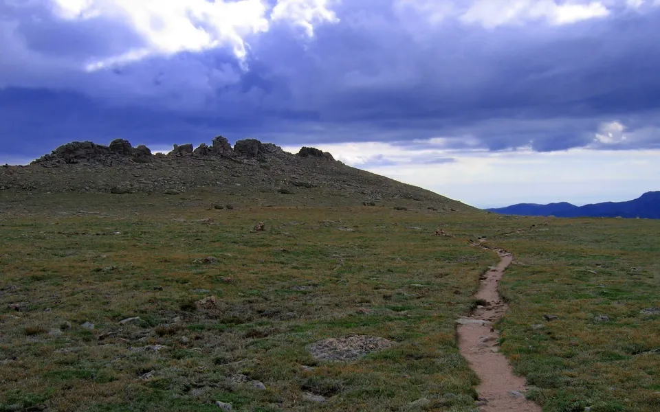 A dirt path winds through a grassy meadow towards a rocky mountain summit under a dramatic cloudy sky.