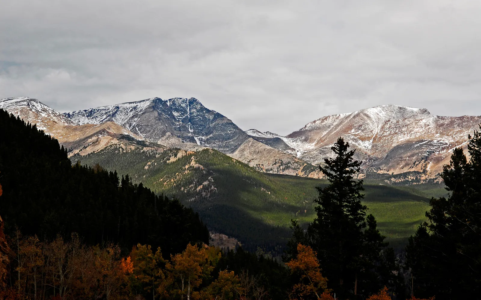 Snow-dusted mountain peaks under a cloudy sky, with dark pine forests and autumn foliage in the foreground.