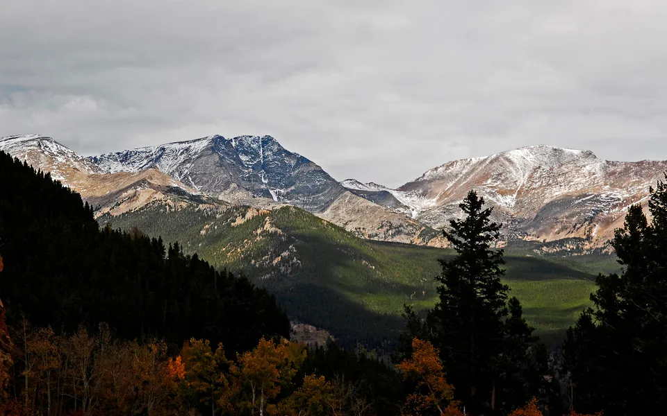 Snow-dusted mountain peaks under a cloudy sky, with dark pine forests and autumn foliage in the foreground.