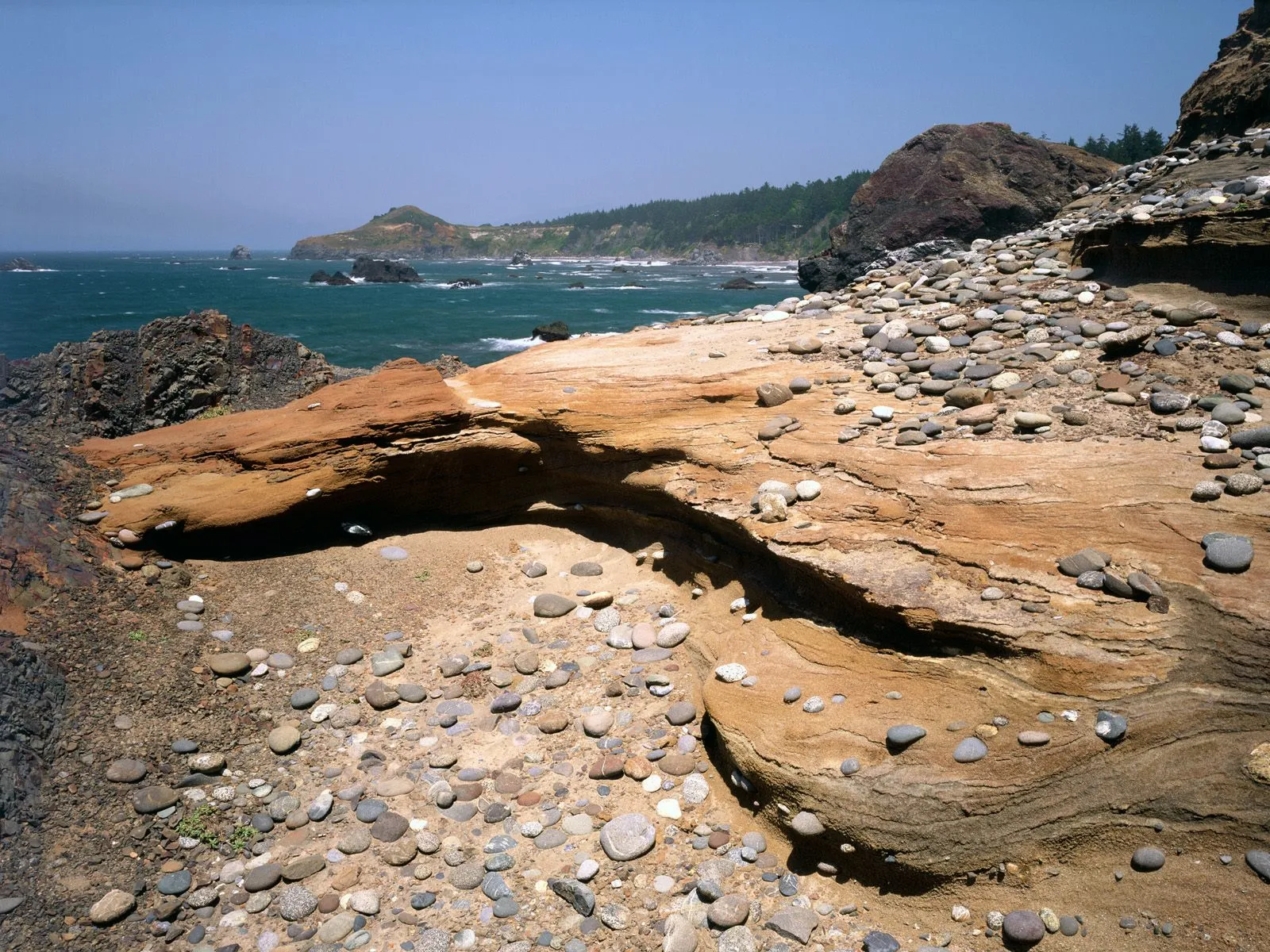 A rugged coastline with weathered orange and gray rocks, pebbles, and the blue ocean under a clear sky.