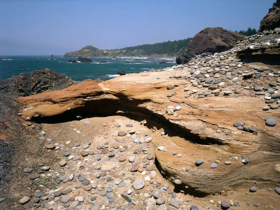 A rugged coastline with weathered orange and gray rocks, pebbles, and the blue ocean under a clear sky.