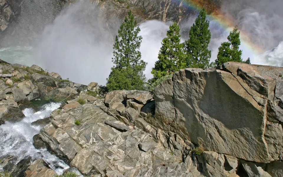 A rainbow appears through the mist above a rocky terrain overlooking a waterfall.