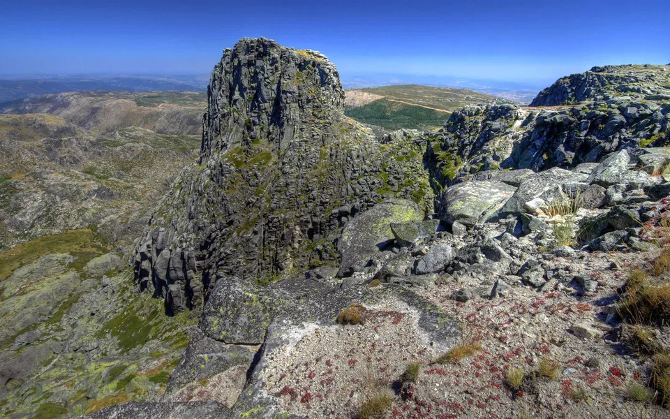 A towering, rugged rock formation dominates the foreground, with a vast, mountainous landscape stretching into the distance under a clear blue sky.