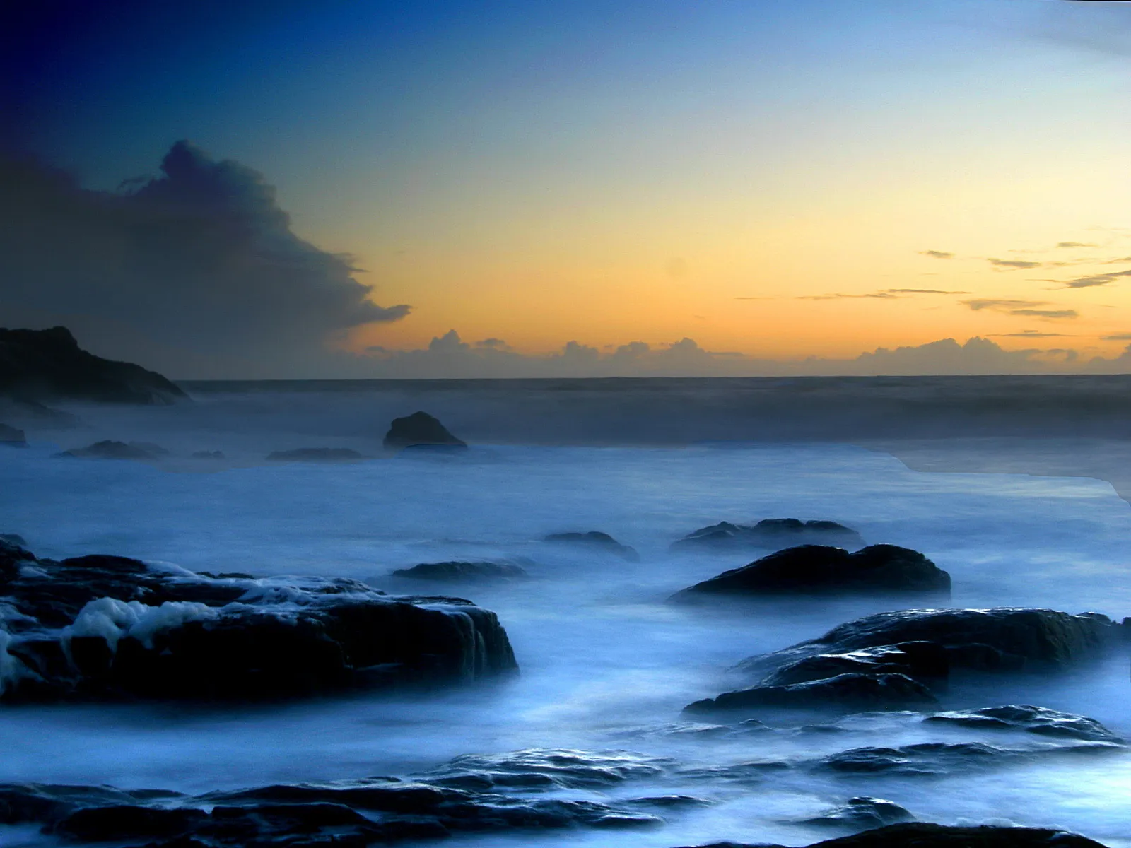 Dark, wet rocks are surrounded by misty, blue ocean water with a soft orange and blue sky in the background.