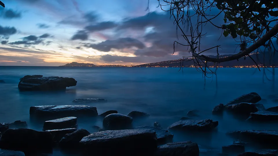 A rocky shoreline with smooth, blurred water under a twilight sky with distant city lights.