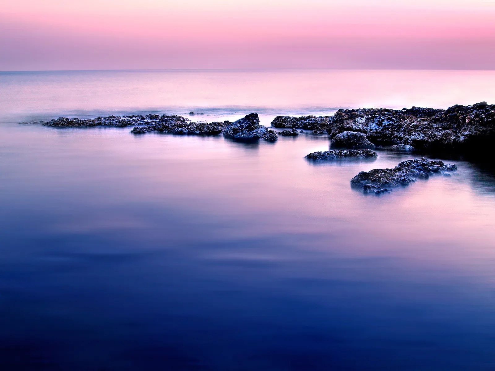 A long exposure shot of a rocky shoreline with smooth water reflecting the pink and purple sky.