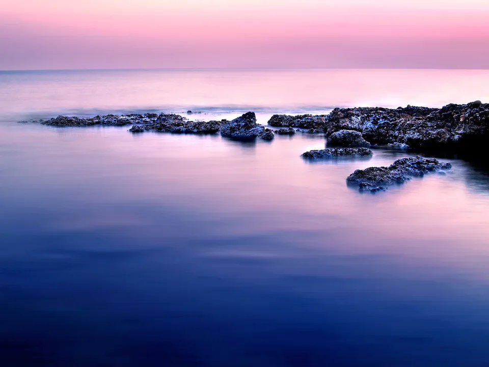 A long exposure shot of a rocky shoreline with smooth water reflecting the pink and purple sky.