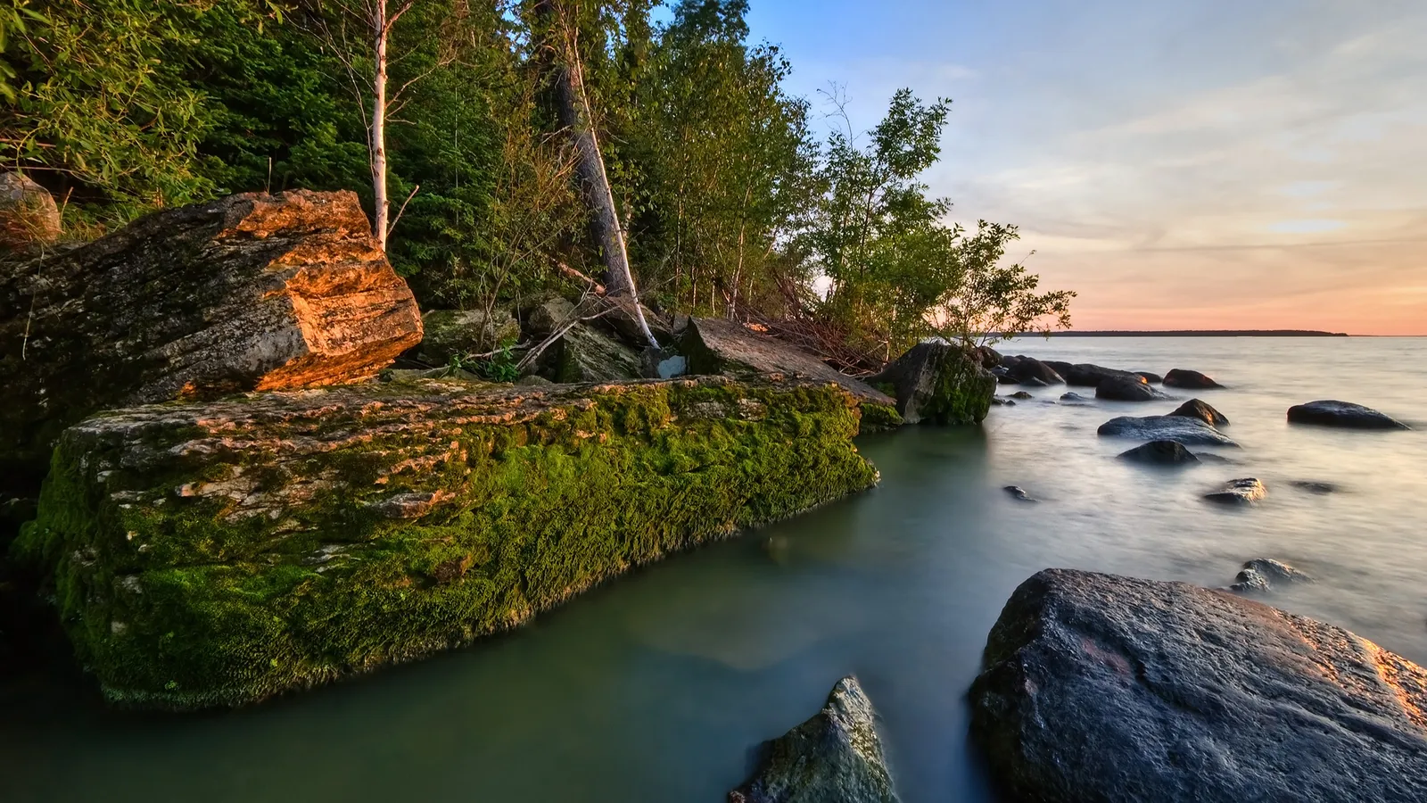 Moss-covered rocks line a calm body of water with a soft sunset sky in the background.