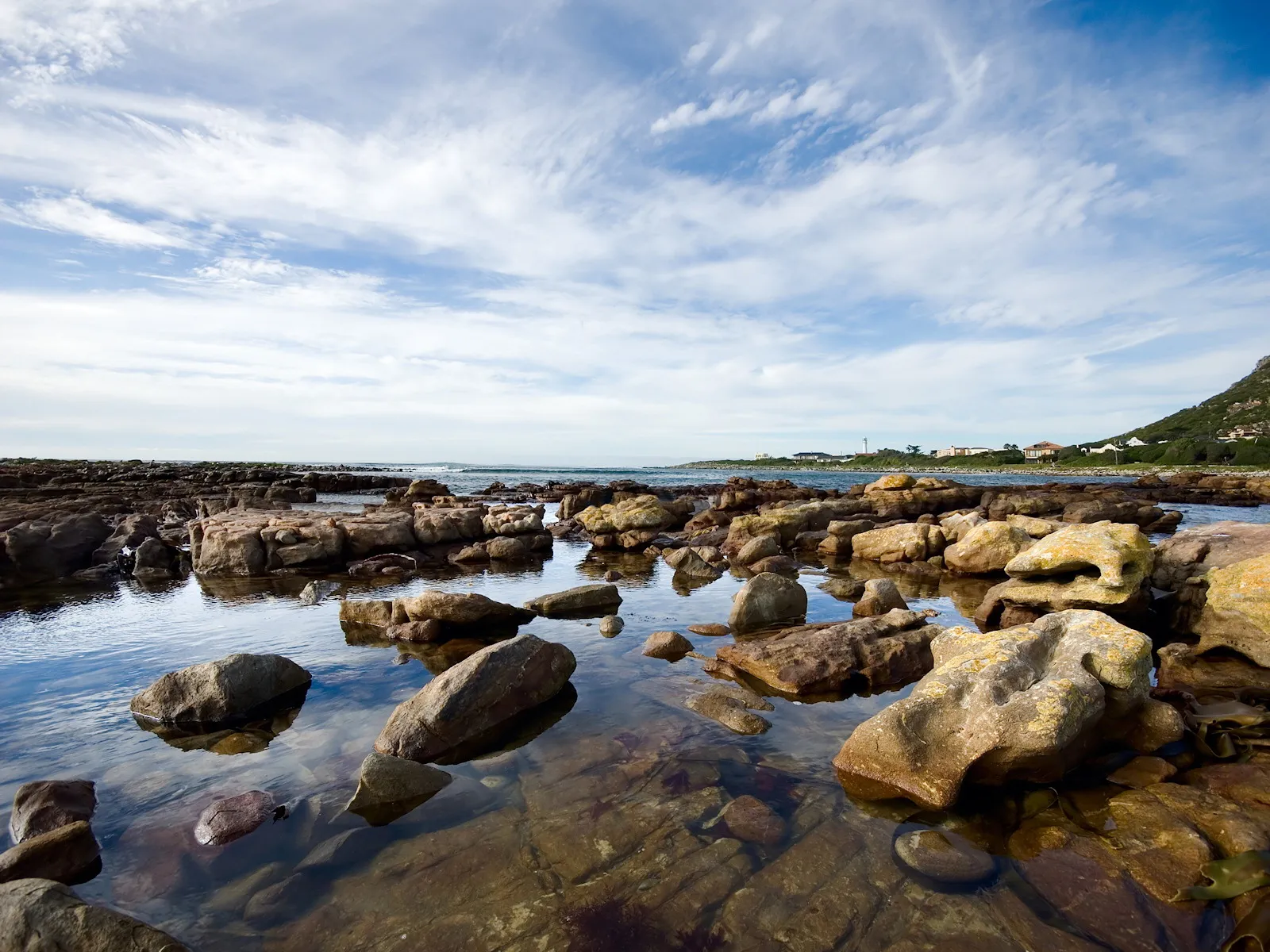A rocky shoreline with shallow tidal pools reflecting the blue sky and scattered clouds.