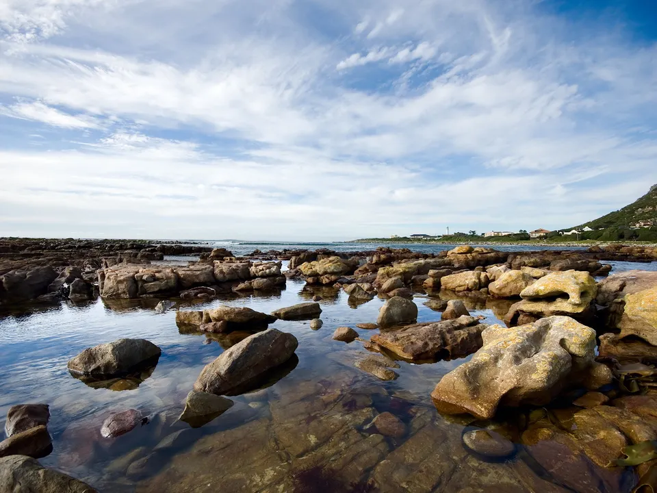 A rocky shoreline with shallow tidal pools reflecting the blue sky and scattered clouds.