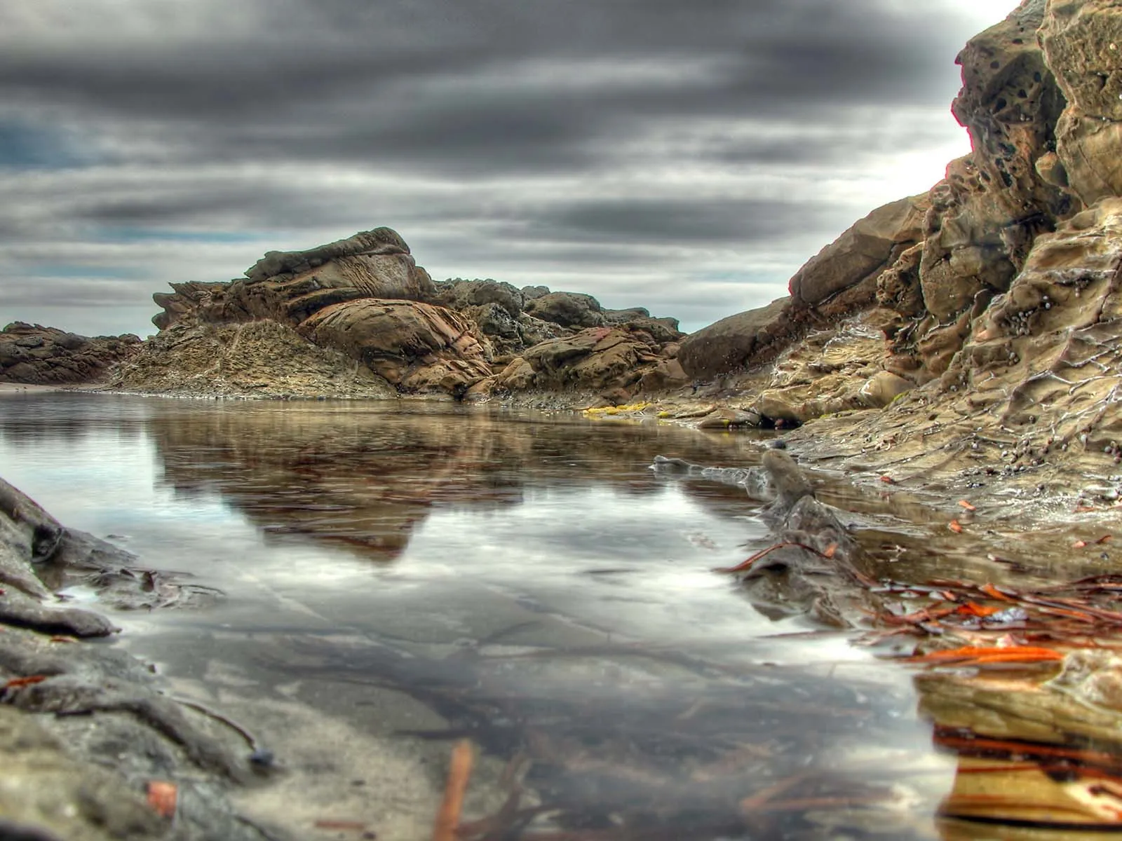 A tide pool reflects dramatic rock formations under a cloudy sky on a rocky shoreline.