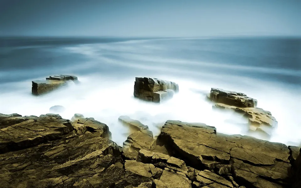 Jagged rocks jut out from a misty shoreline as waves crash around them.