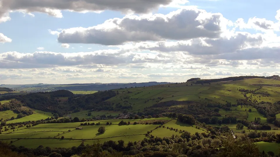 A wide view of green rolling hills with scattered trees and a village in the distance under a partly cloudy sky.