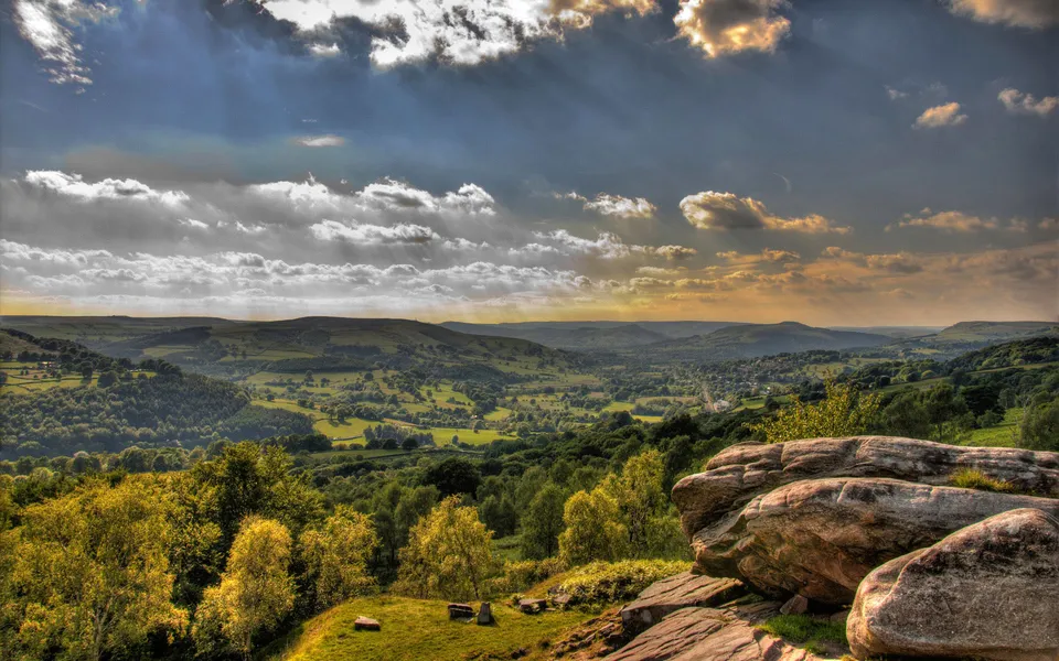 A wide vista of green rolling hills and valleys under a dramatic sky with sunbeams breaking through clouds.