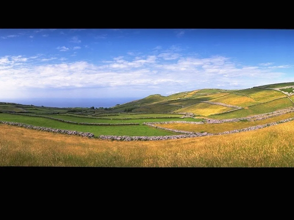 A panoramic view of green rolling hills with stone walls dividing fields under a blue sky with scattered clouds.