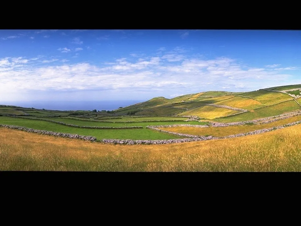 A panoramic view of green rolling hills with stone walls dividing fields under a blue sky with scattered clouds.