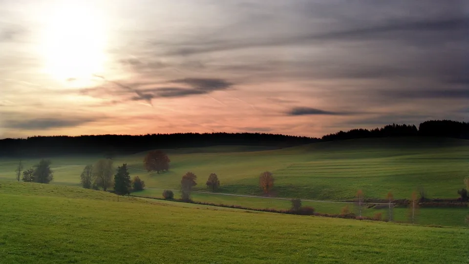 A wide shot of rolling green hills under a dramatic sky at sunset, with trees scattered across the landscape.