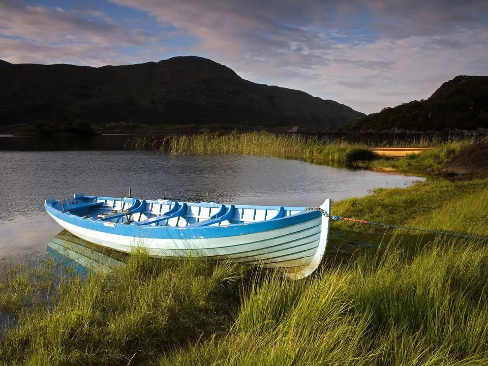 A blue and white rowboat is tied to the grassy shore of a calm lake, with dark hills in the background.