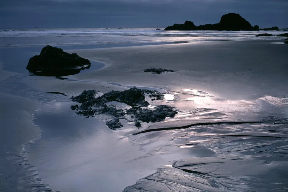 A dark, moody scene of wet sand with tide pools reflecting the sky and rocks at Ruby Beach.