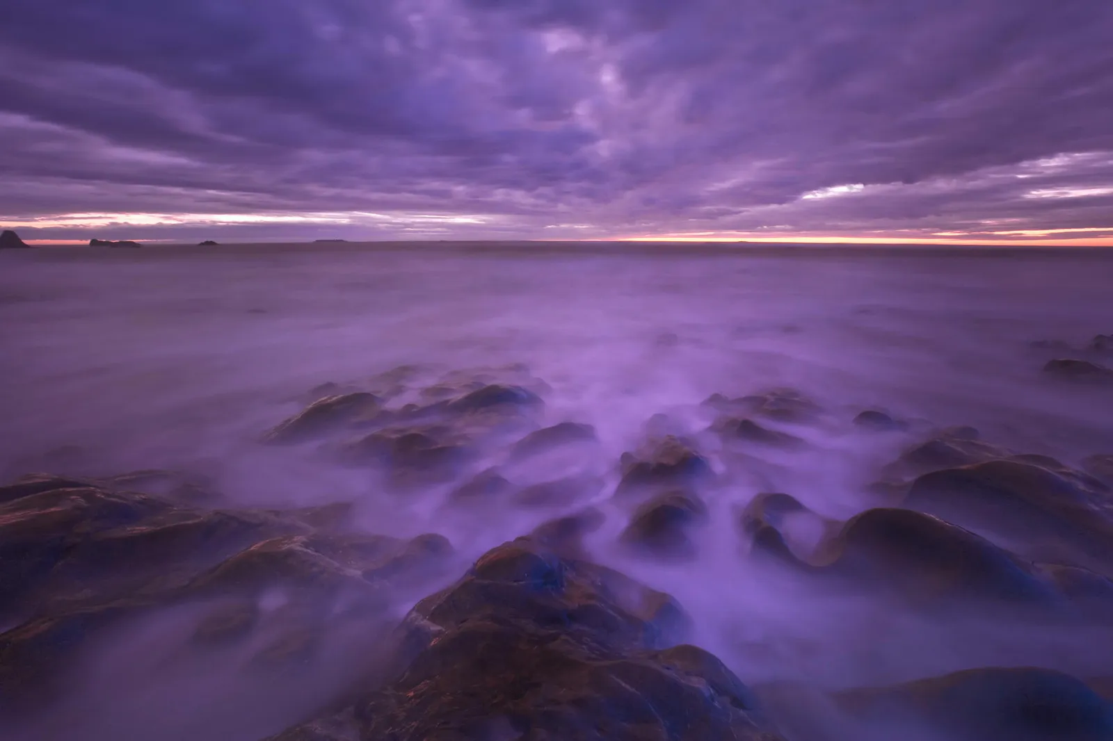 A misty, purple-hued sunset over a rocky shoreline with the ocean waves creating a soft, ethereal effect.