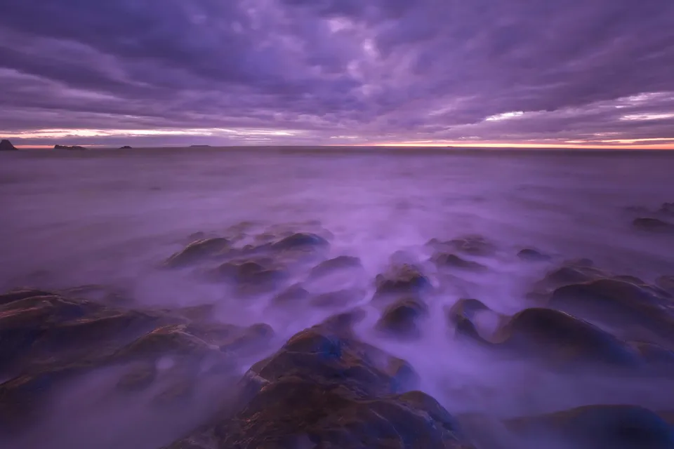 A misty, purple-hued sunset over a rocky shoreline with the ocean waves creating a soft, ethereal effect.