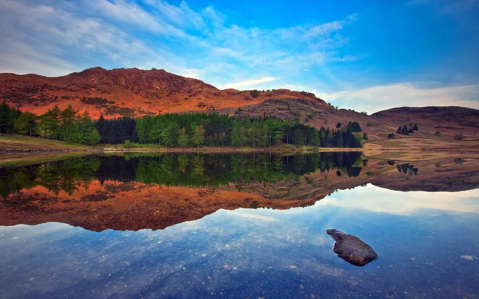 Rugged, orange-hued mountains are mirrored in the still waters of a lake.