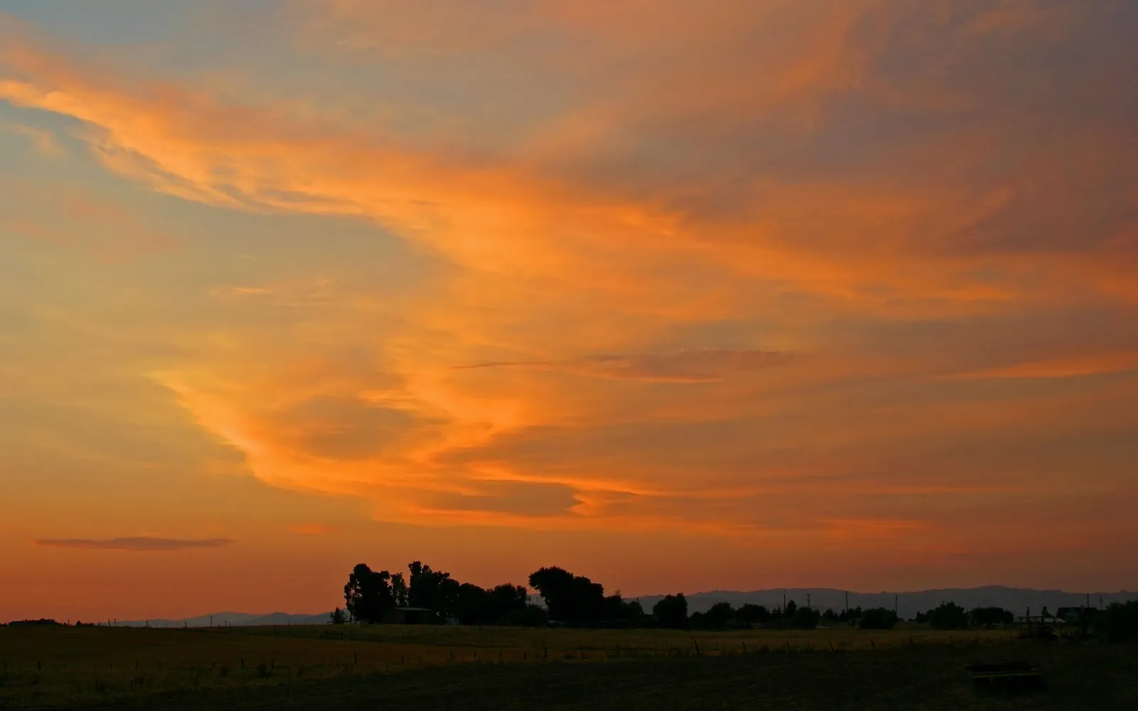 A vibrant orange and yellow sky with wispy clouds over a dark silhouette of trees and hills at sunset.