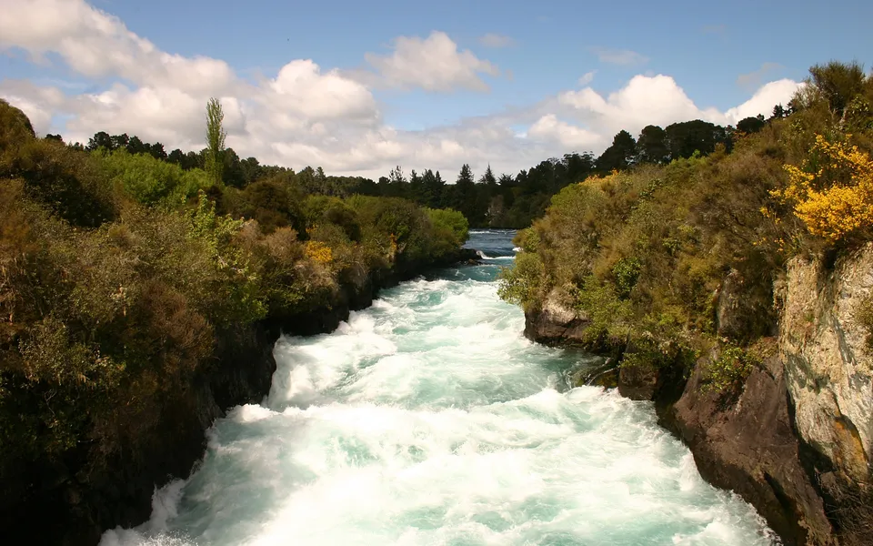 A wide, turbulent river with white water rapids flows between rocky, tree-covered banks under a blue sky with white clouds.