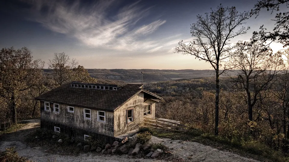A weathered wooden cabin sits on a hill overlooking a vast, tree-covered valley under a dramatic sky.