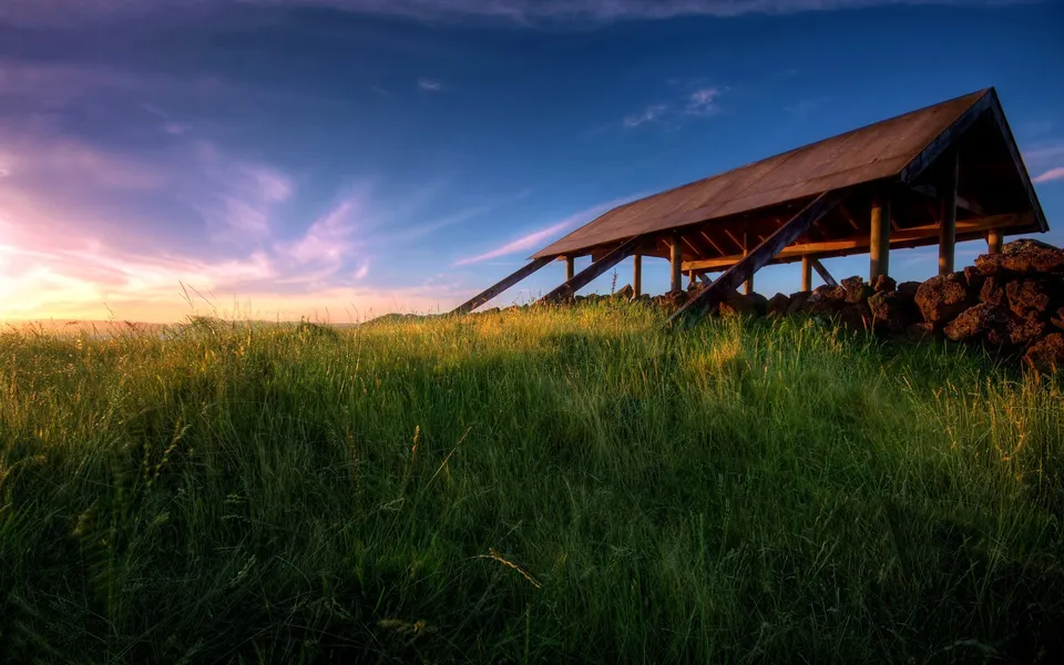 A wooden open shelter with a rustic stone wall sits on a grassy hill at sunset.