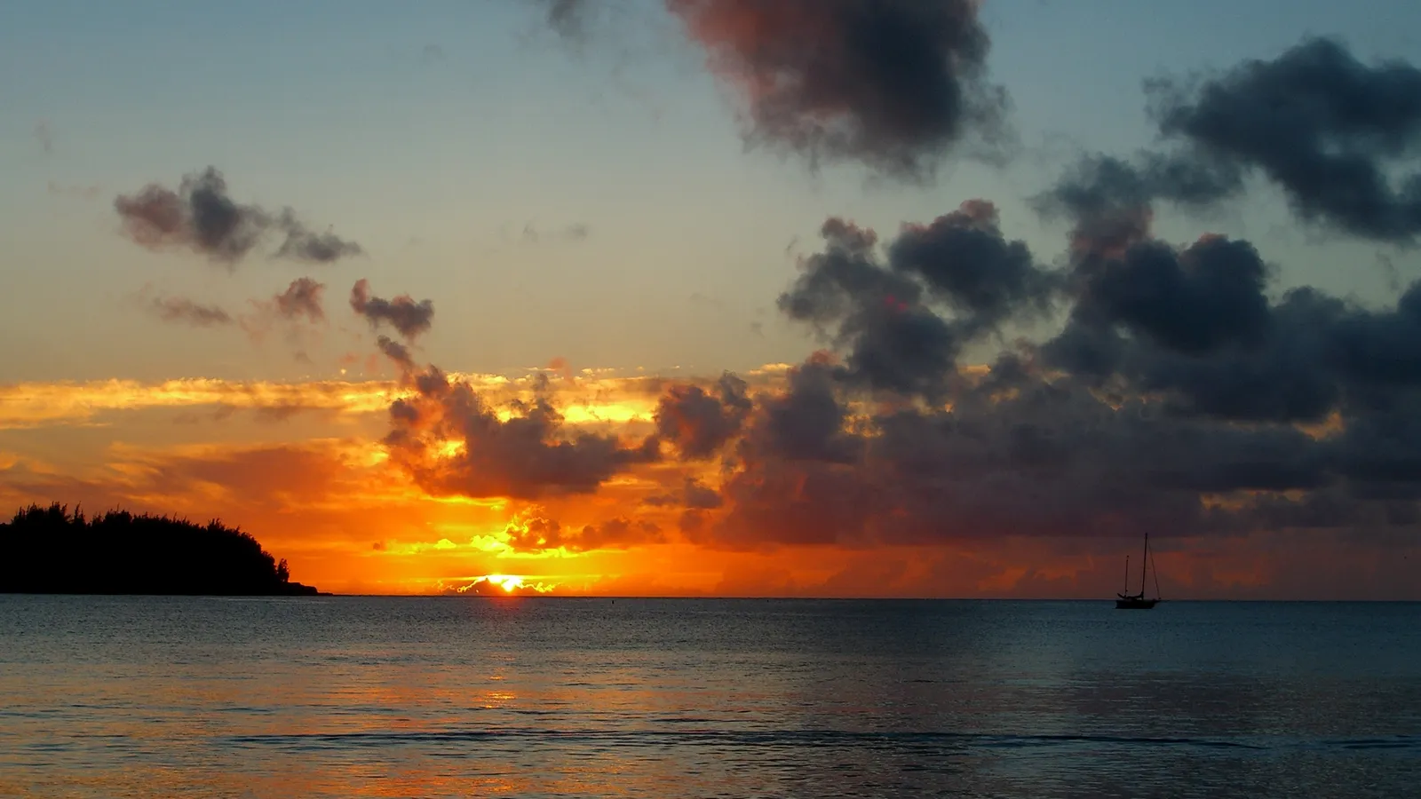A sailboat is silhouetted against a dramatic sunset with orange and yellow clouds over the ocean.