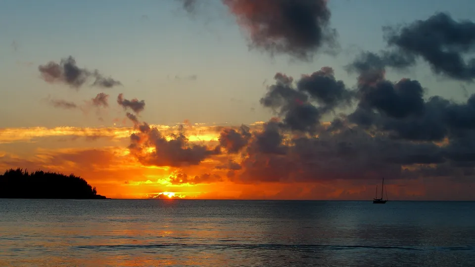 A sailboat is silhouetted against a dramatic sunset with orange and yellow clouds over the ocean.