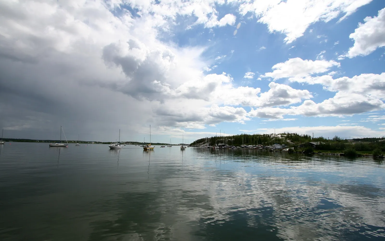 Sailboats are moored in a calm lake with a shoreline of trees and buildings under a dramatic sky with both dark storm clouds and bright white clouds.