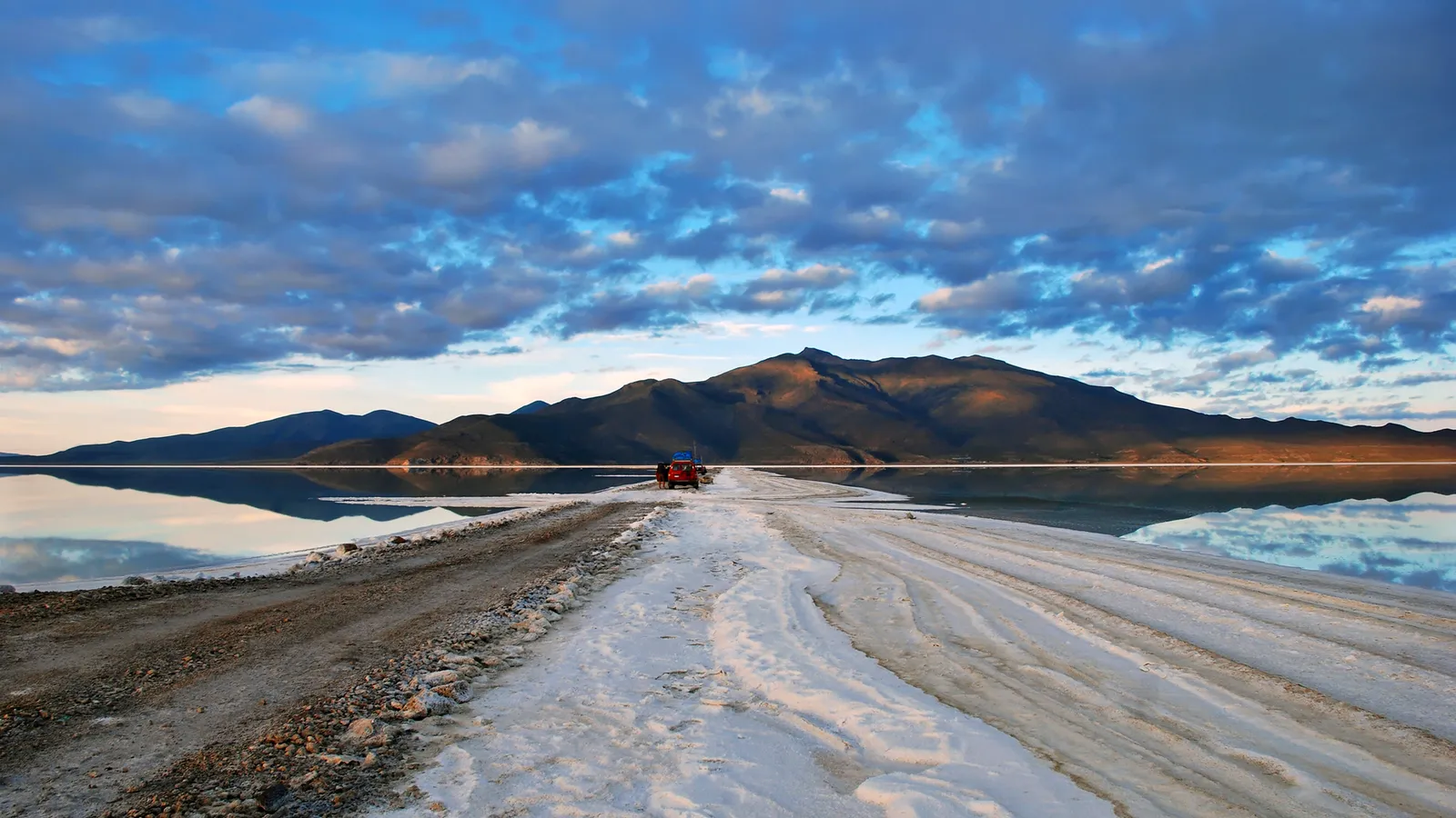A red car drives on a dirt and salt road that leads across a vast, reflective salt flat towards distant mountains under a cloudy blue sky.