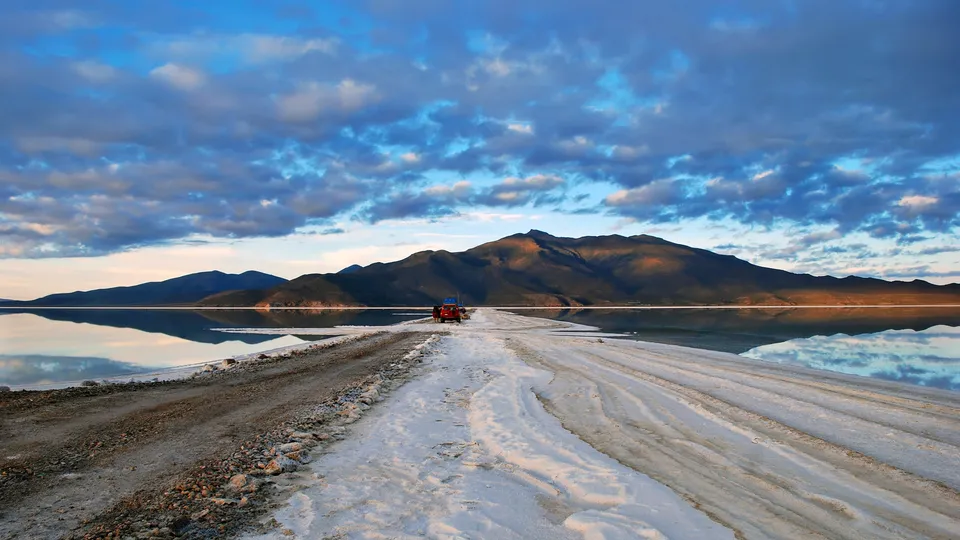 A red car drives on a dirt and salt road that leads across a vast, reflective salt flat towards distant mountains under a cloudy blue sky.