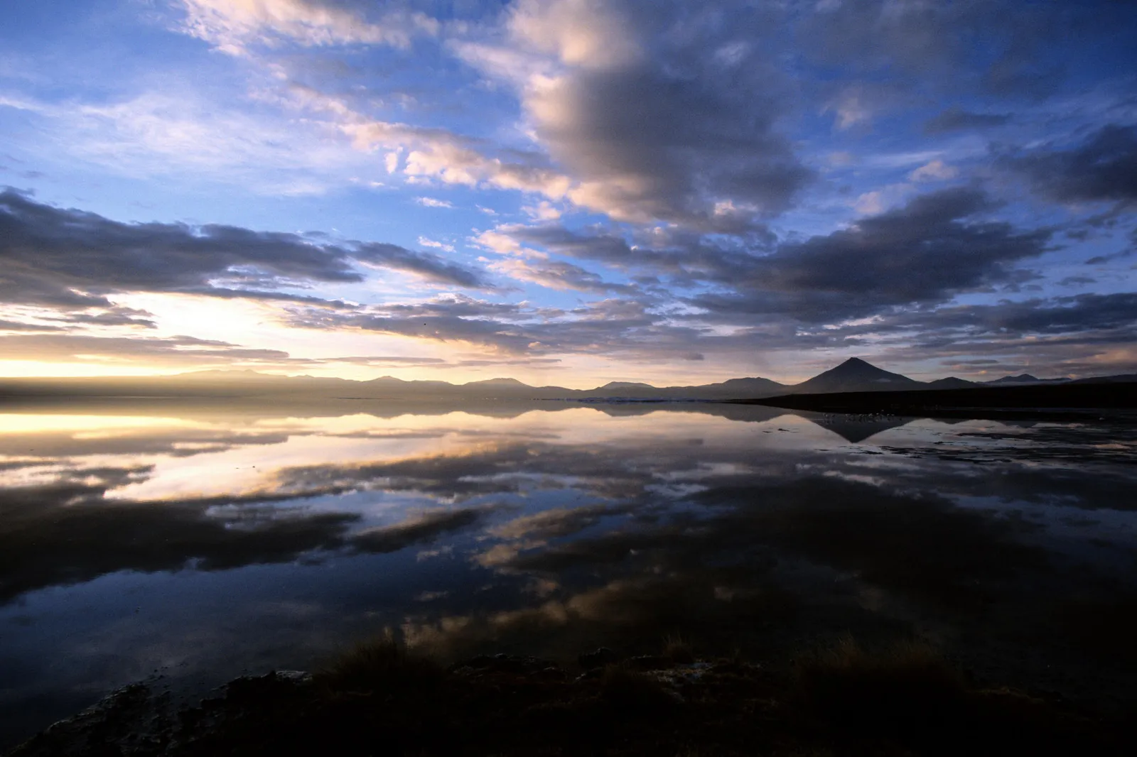 A vast salt lake reflects the dramatic clouds and distant mountains during sunset.