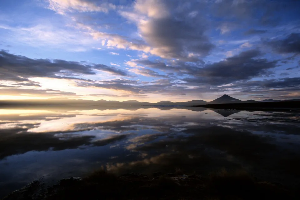 A vast salt lake reflects the dramatic clouds and distant mountains during sunset.