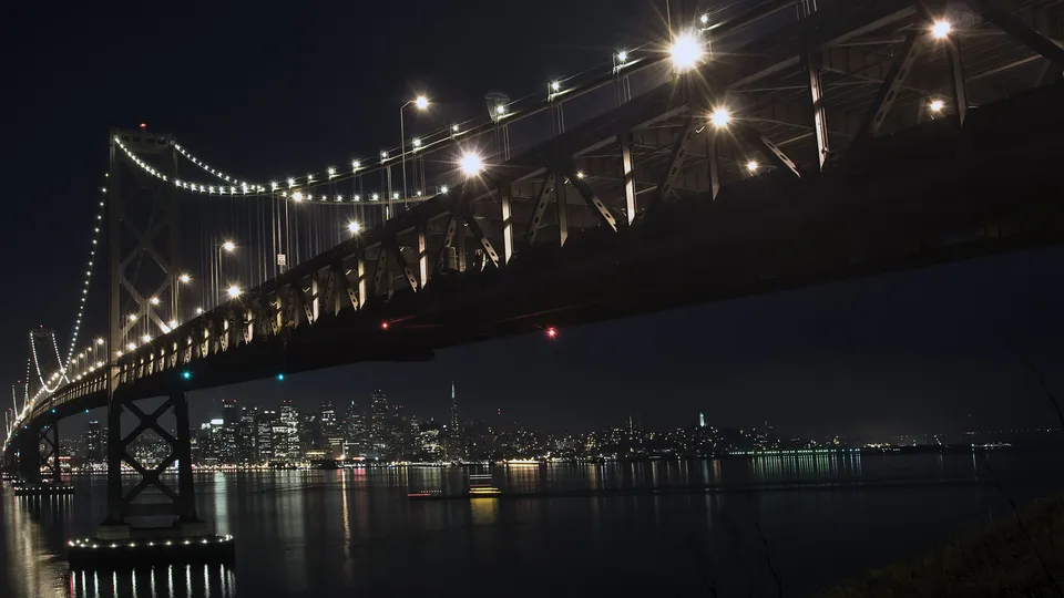 The illuminated San Francisco Bay Bridge spans across dark water with the city skyline in the background at night.
