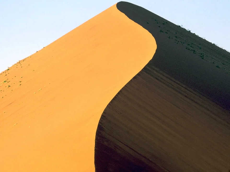 A large sand dune with a sharp curve, casting a shadow on one side and illuminated by sunlight on the other.