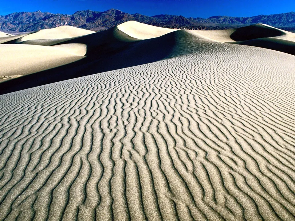 Vast sand dunes with ripples are set against distant mountains and a blue sky.