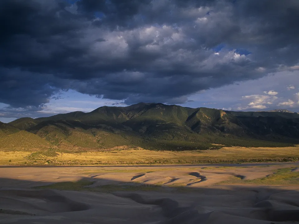 Sand dunes in the foreground with mountains and dramatic clouds in the background.