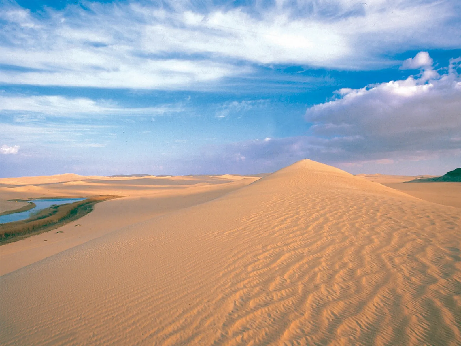 Expansive sand dunes stretch towards the horizon under a blue sky with scattered clouds, with a small body of water visible to the left.