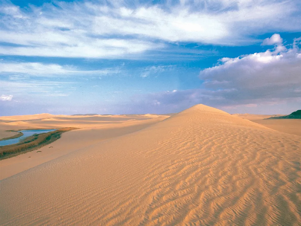 Expansive sand dunes stretch towards the horizon under a blue sky with scattered clouds, with a small body of water visible to the left.