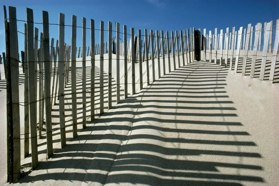 A wooden sand fence casts long, striped shadows across a sandy dune under a clear blue sky.