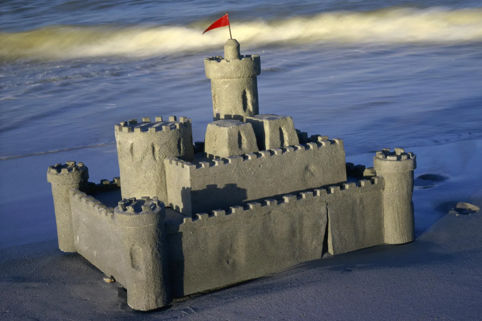 A detailed sandcastle with towers and walls sits on a sandy beach, with gentle waves in the background.